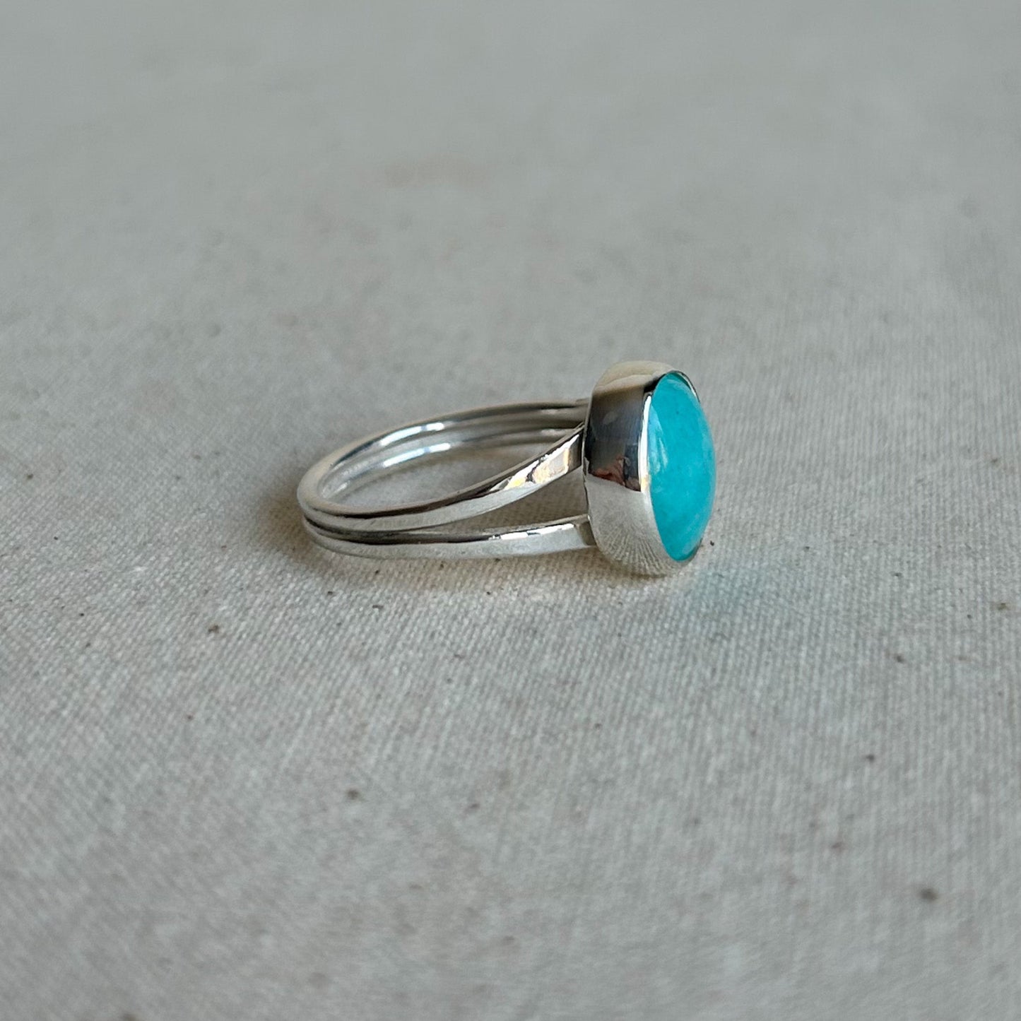 A silver ring with a round turquoise cabochon gemstone, resting on a textured light beige fabric surface.