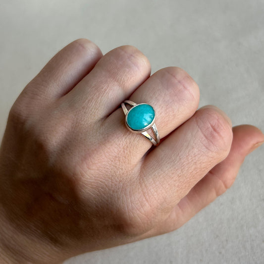 A silver ring with an oval turquoise cabochon stone worn on a person's ring finger, shown against a neutral light gray background.