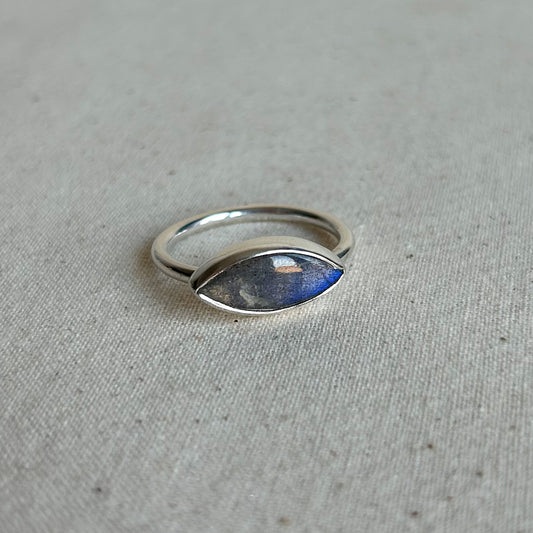 A silver ring with a marquise-shaped blue stone, photographed on a textured beige fabric background.
