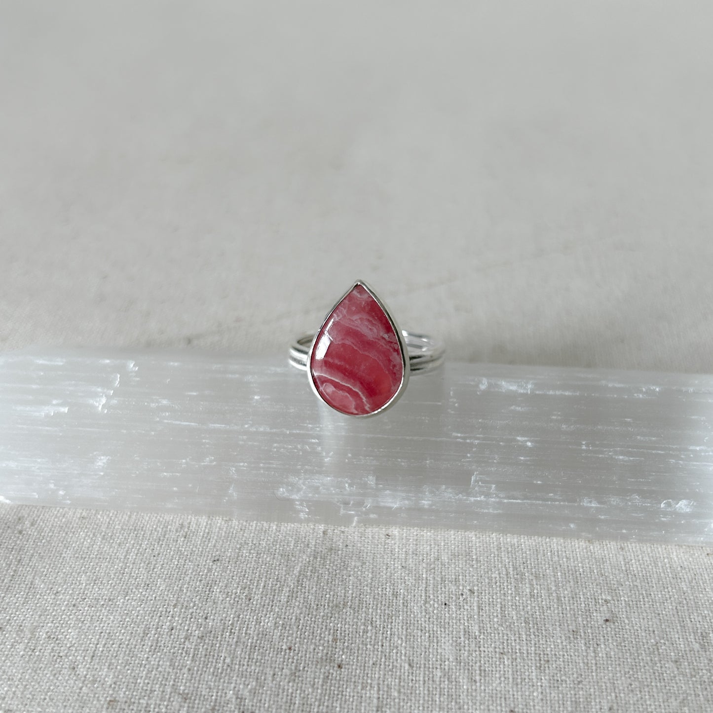 A silver teardrop-shaped rhodochrosite ing with a pink  gemstone set in it, placed on a white surface