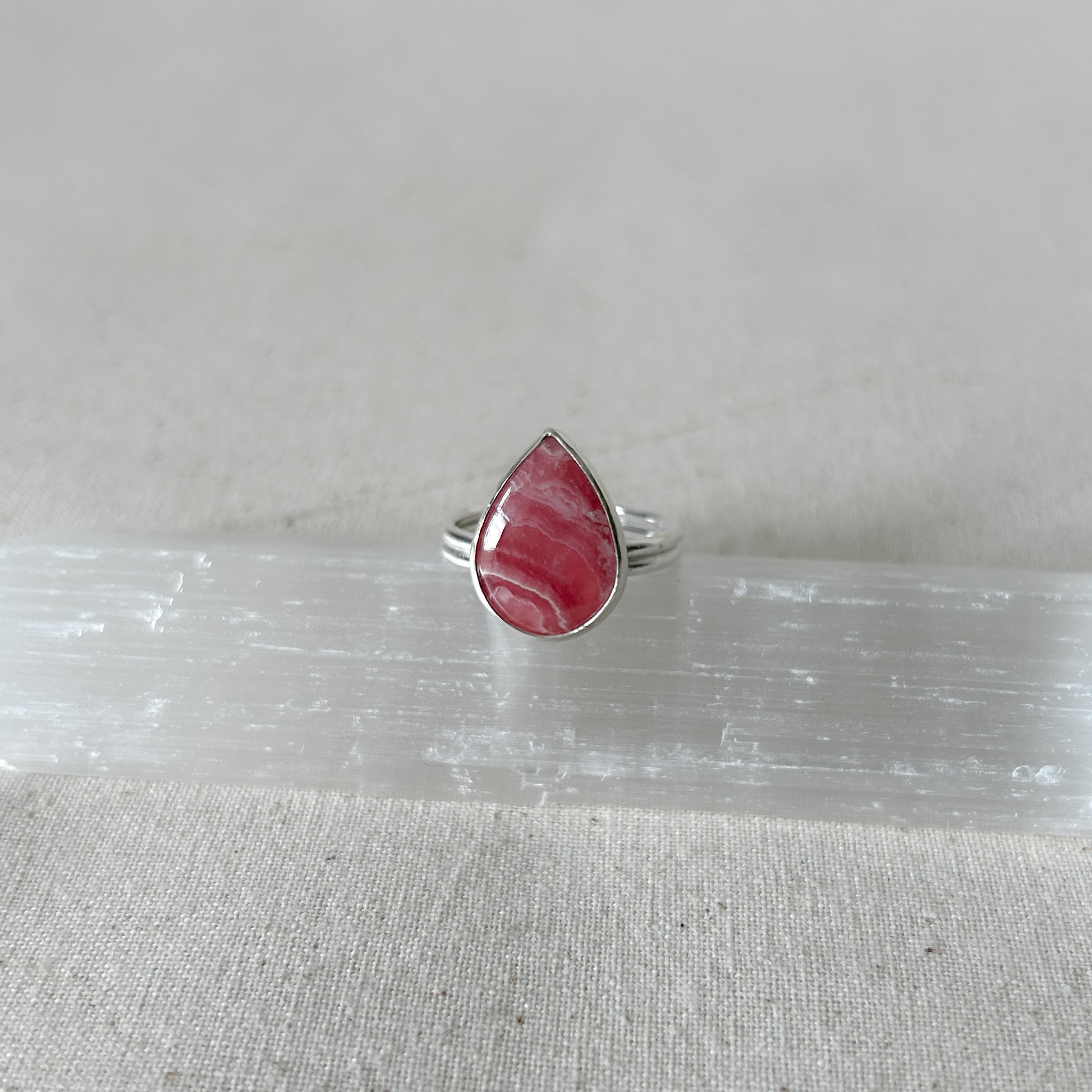A silver teardrop-shaped rhodochrosite ing with a pink  gemstone set in it, placed on a white surface