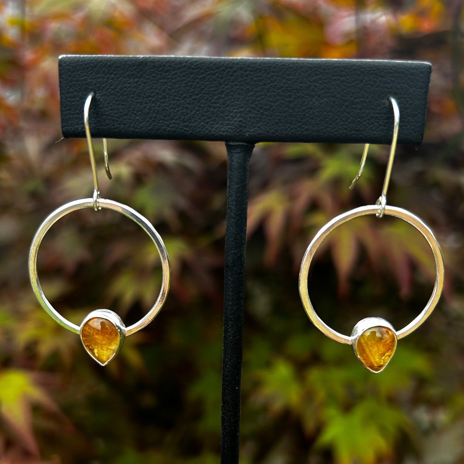 Silver dangle earrings with honey coloured stones on a black stand against a blurred natural background