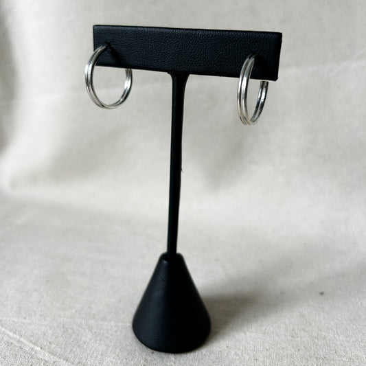 A pair of silver huggie hoops  displayed on a black stand with a textured surface.