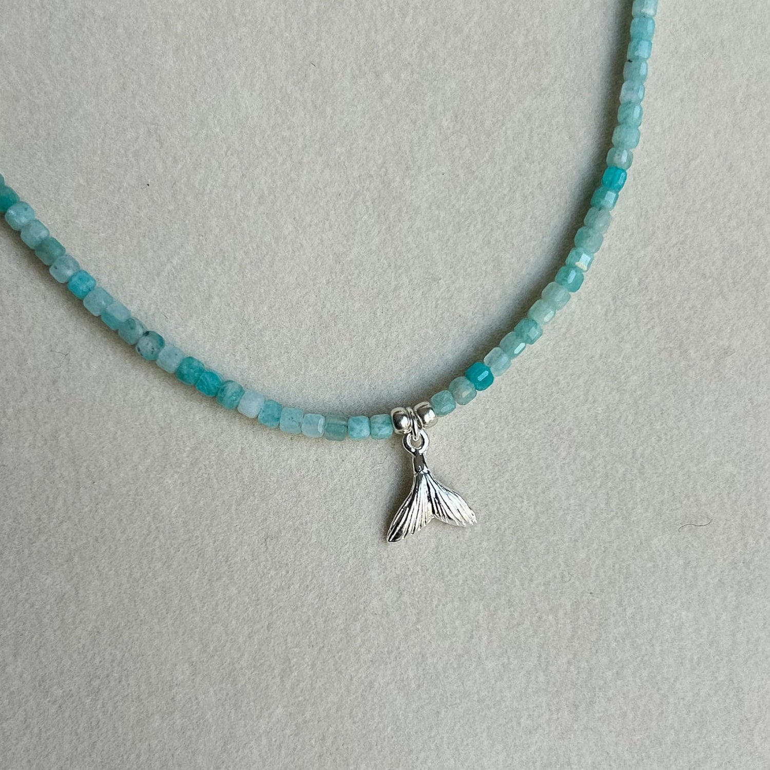 A beaded necklace with faceted turquoise-blue beads and a small silver mermaid tail pendant, displayed on a light gray jewelry bust.