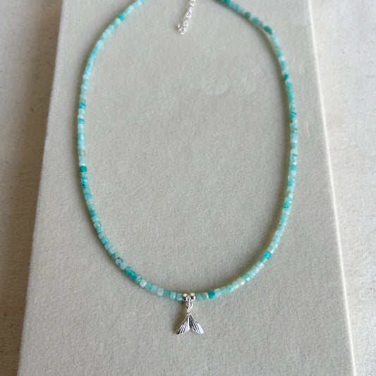 A beaded necklace with small turquoise-blue and white beads and a silver mermaid tail charm, displayed on a light gray jewelry box.