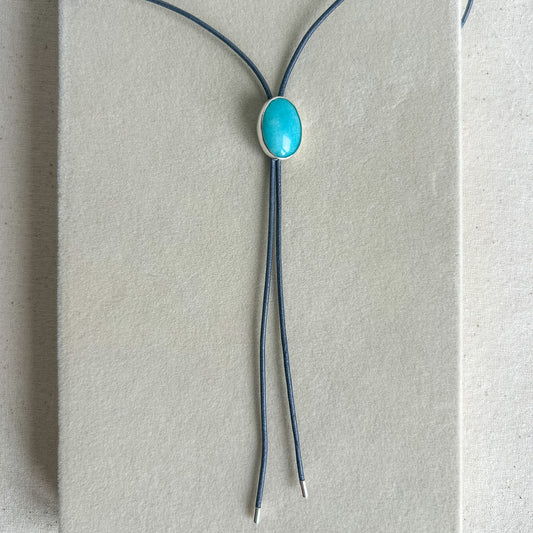 A bolo-style necklace with a smooth oval turquoise cabochon in a silver bezel, suspended from two dark blue-gray cords on a textured light beige display card.