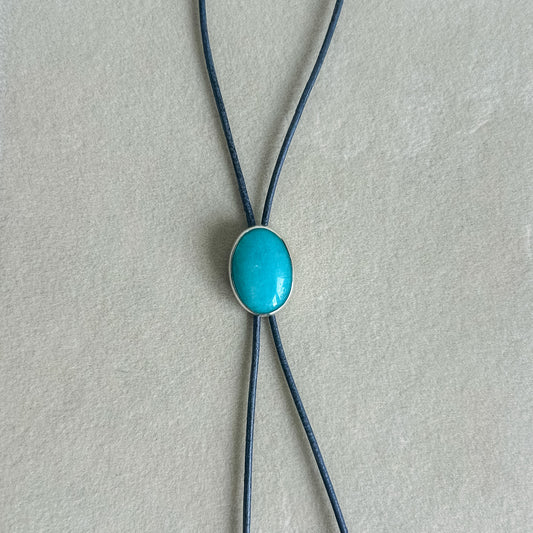 A bolo-style necklace featuring a smooth, oval turquoise cabochon set in a silver-tone bezel, suspended from two dark navy blue cords on a textured light gray background.