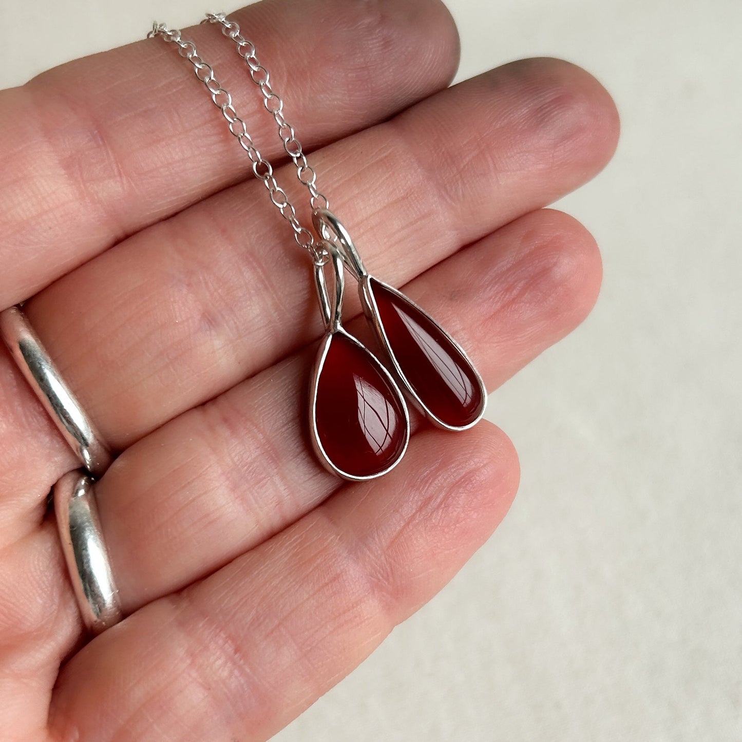 Hand holding a silver necklace with a red teardrop pendant against a plain background