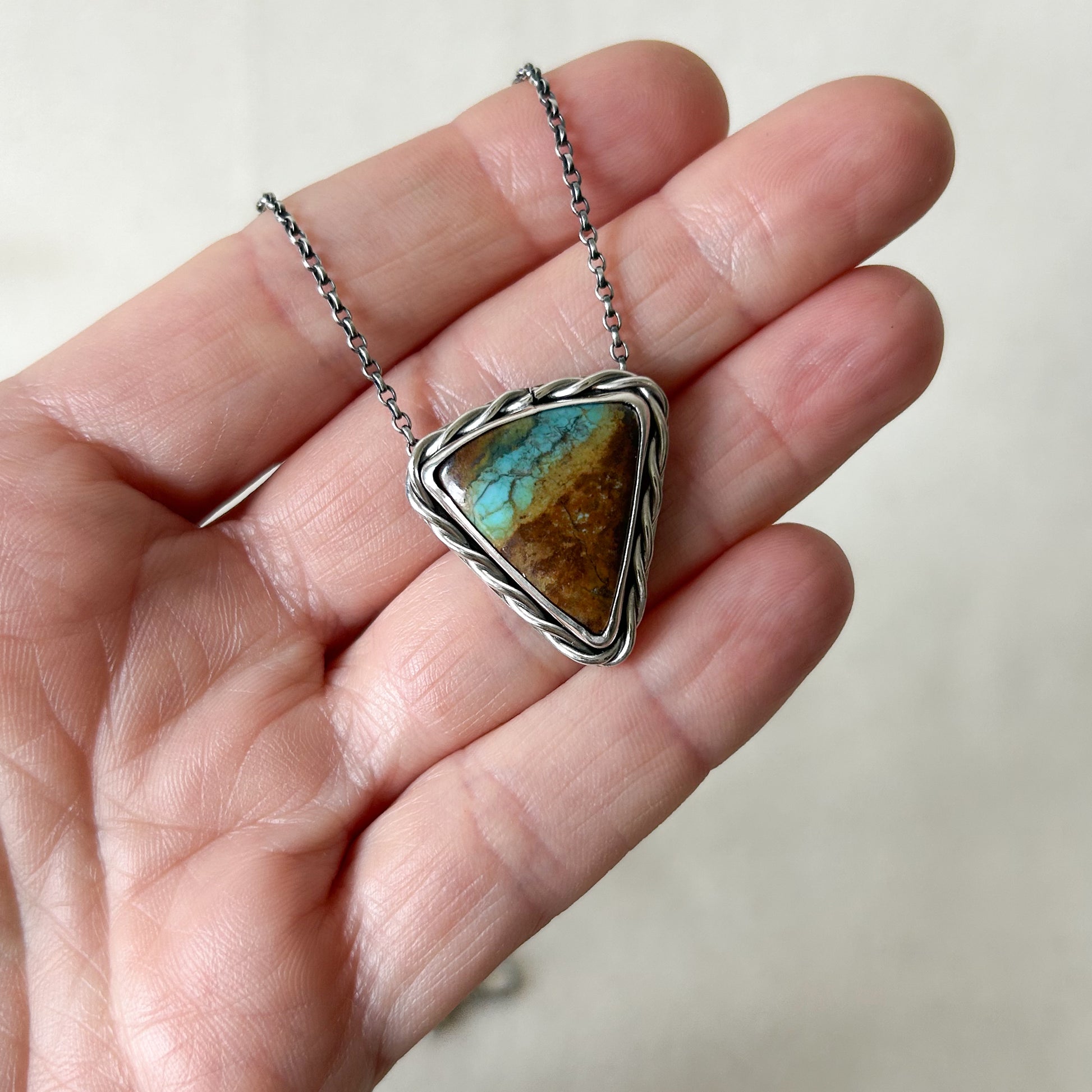 Turquoise stone pendant on a silver chain held in a hand against a neutral background
