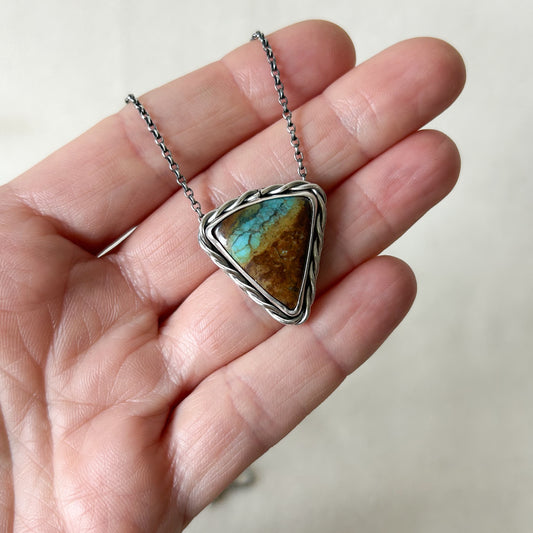 Turquoise stone pendant on a silver chain held in a hand against a neutral background