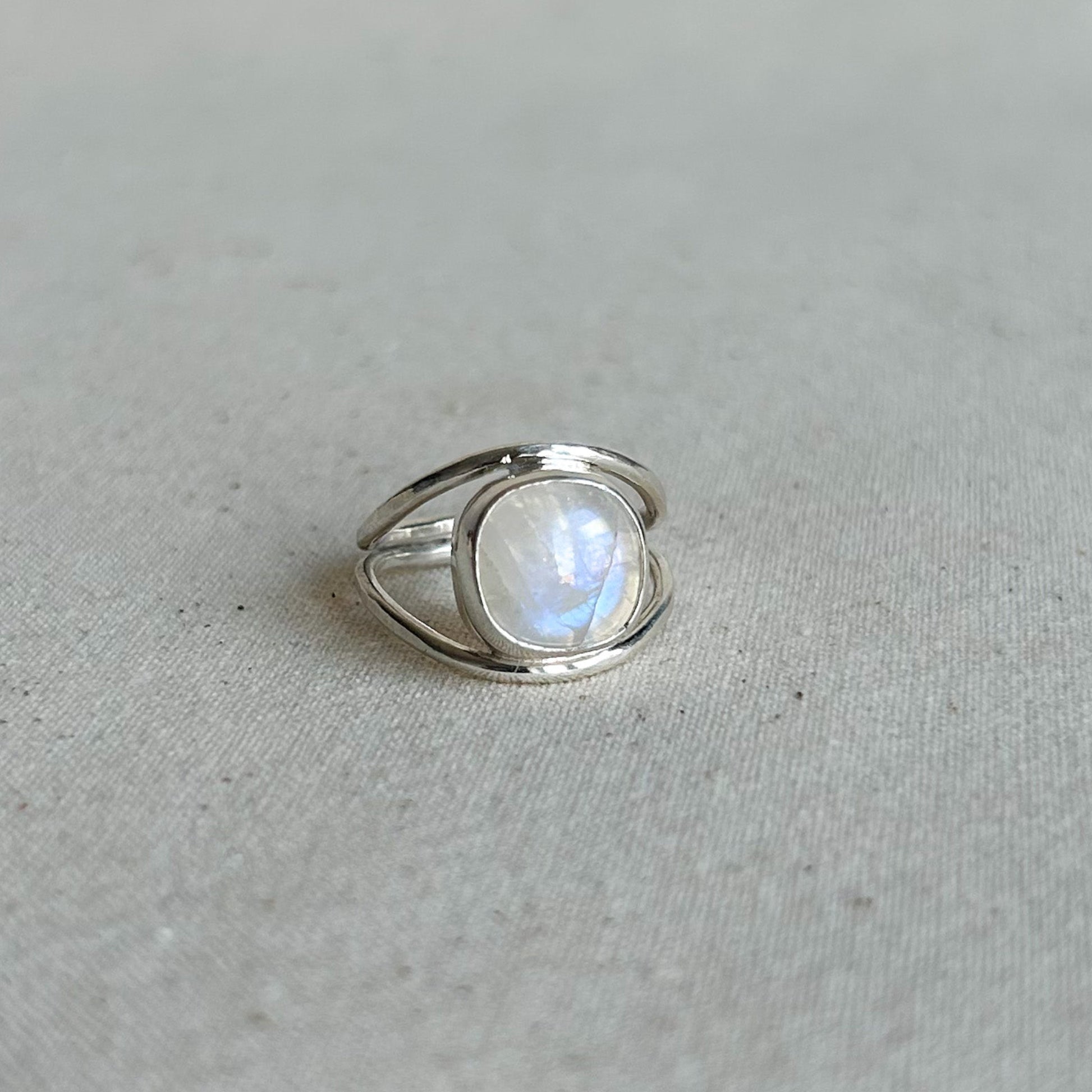A silver ring featuring a large, oval-shaped cabochon moonstone with a soft blue and white iridescent glow, set against a neutral, textured light gray fabric background.