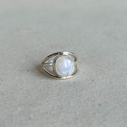 A silver ring featuring a large, oval-shaped cabochon moonstone with a soft blue and white iridescent glow, set against a neutral, textured light gray fabric background.