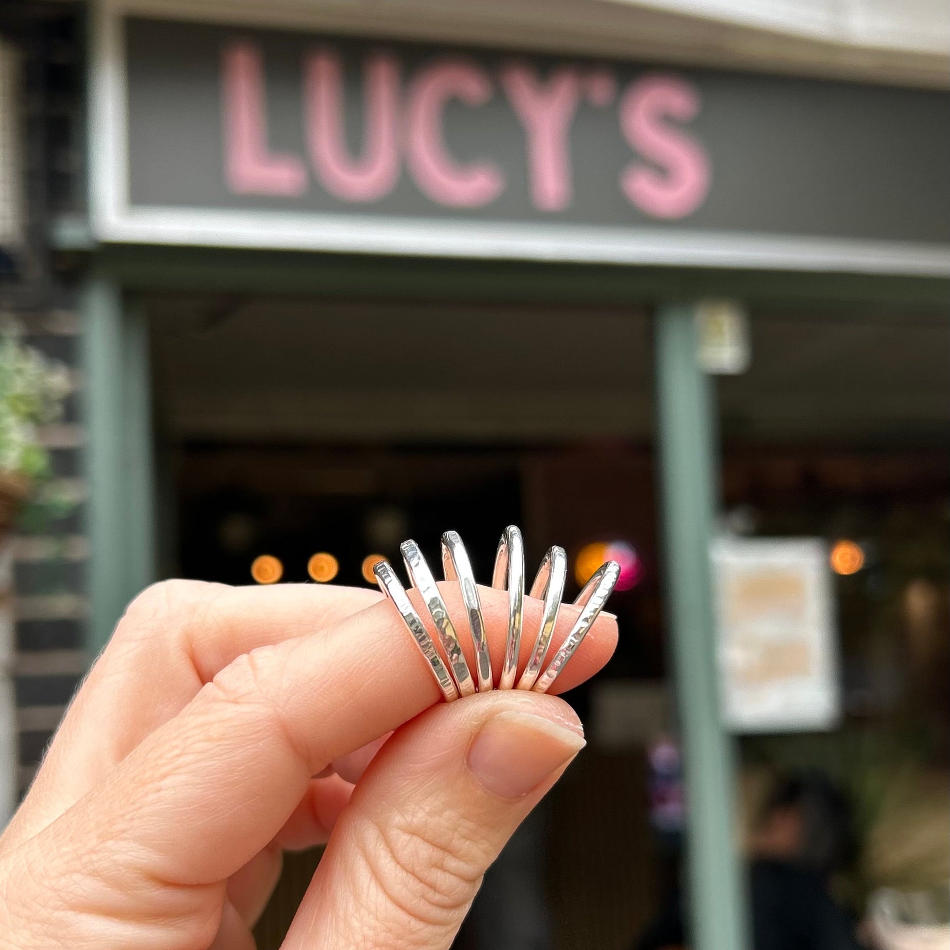 A hand holding a set of five slim silver rings with engraved names, photographed in front of a blurred storefront with a pink 'LUCY'S' sign.