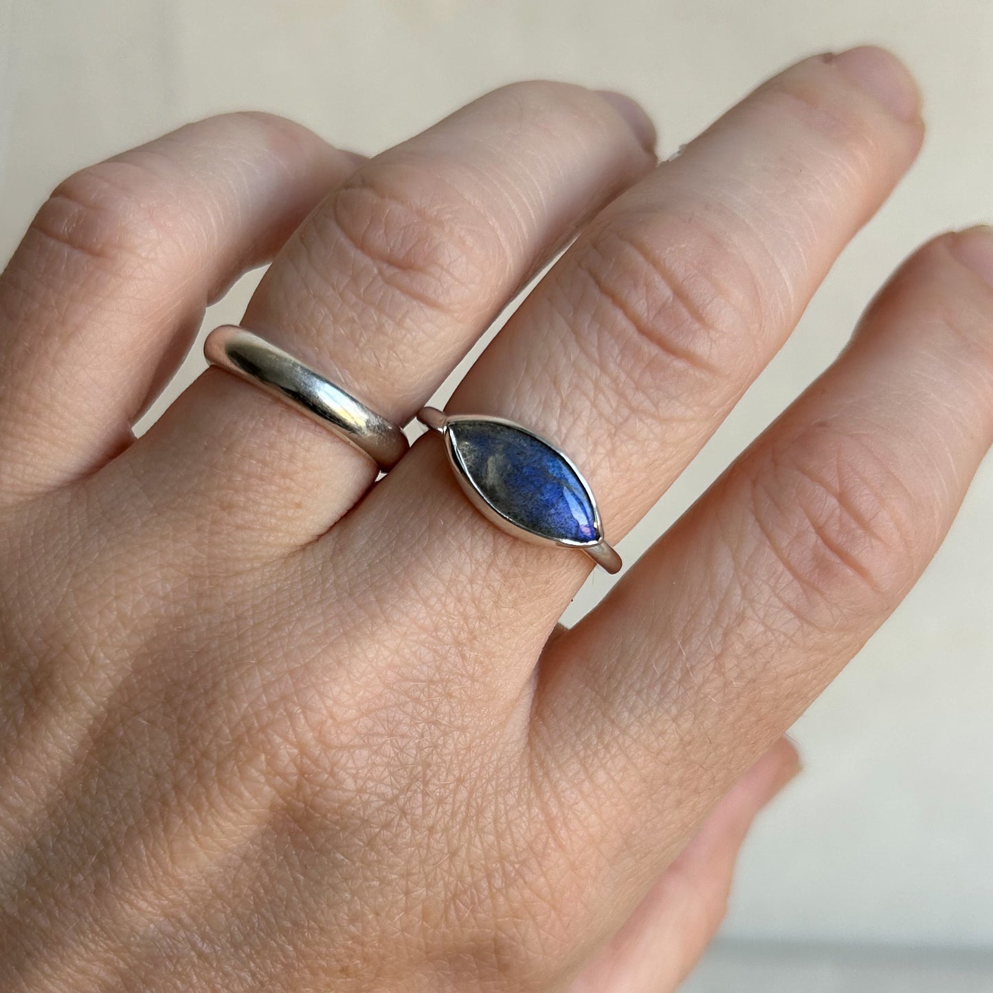 A close-up of a hand wearing two rings: a simple polished silver band on one finger and a silver ring with a marquise-shaped blue lapis lazuli stone on the other finger.