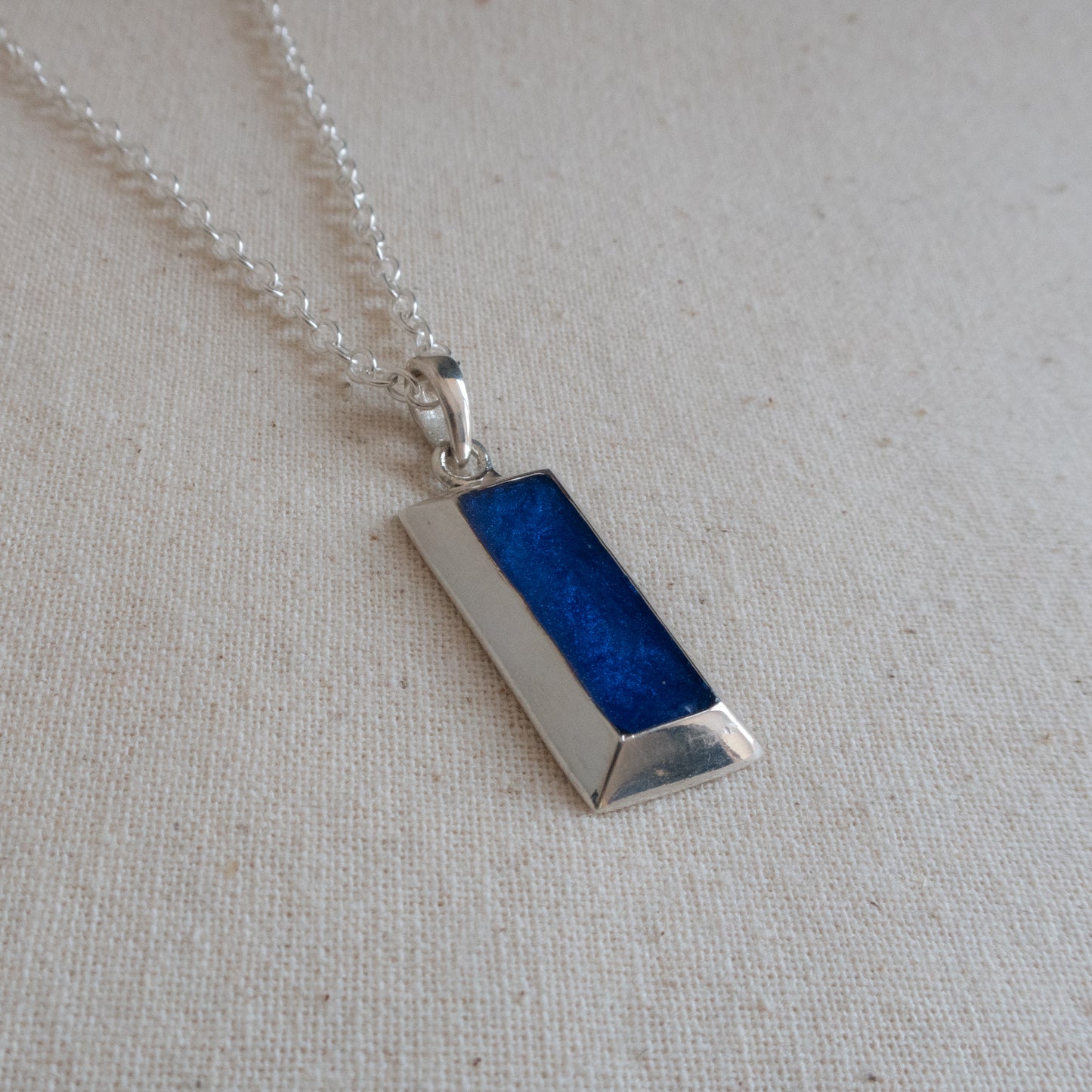 A silver-toned rectangular pendant necklace with a textured, glittery deep blue center panel, resting on a neutral beige linen fabric background.