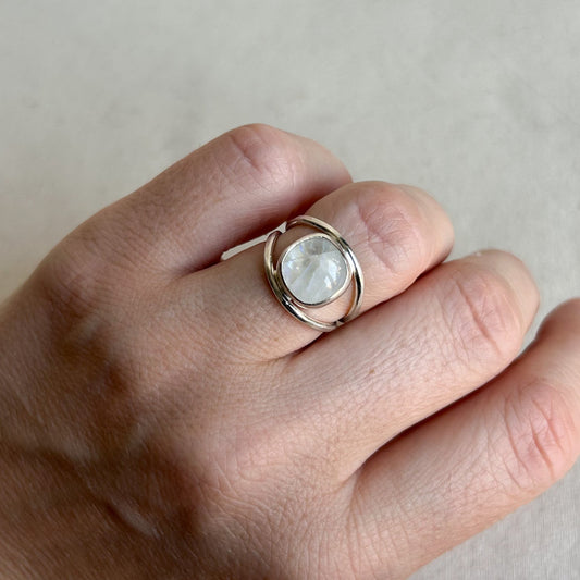 A silver ring with a round, milky white stone set on a hand, shown against a neutral light gray background.