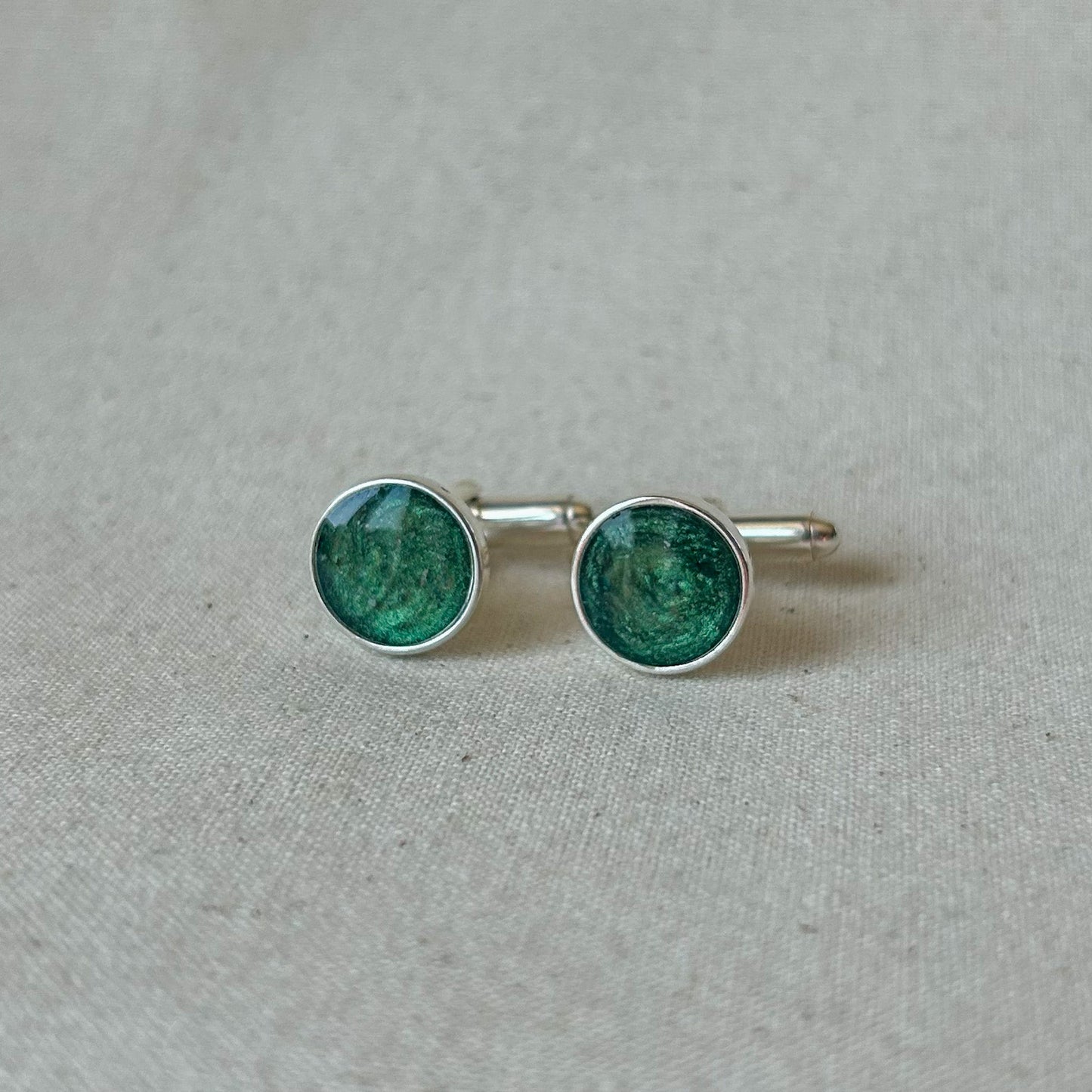 A pair of silver cufflinks with round green enamel inserts, displayed on a textured beige fabric background.