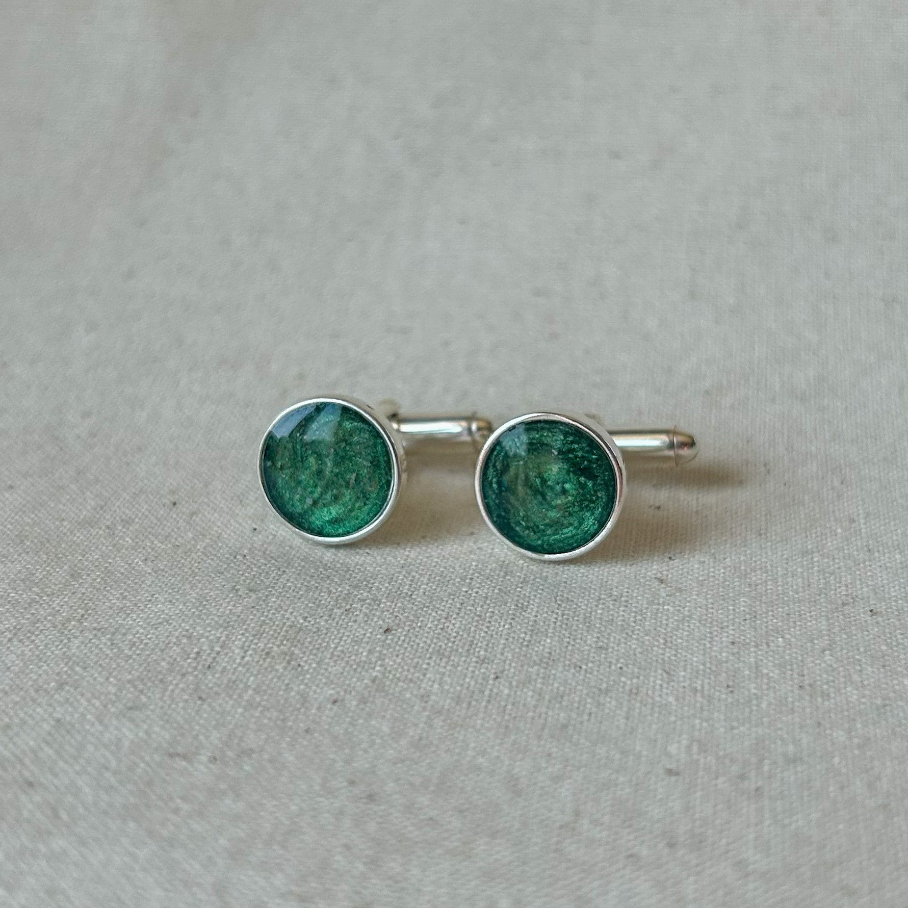 A pair of silver cufflinks with round green enamel inserts, displayed on a textured beige fabric background.