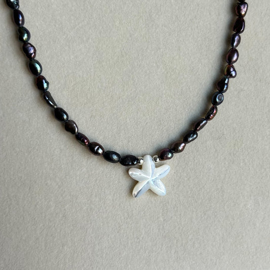A beaded necklace with dark, iridescent baroque pearls and a small white starfish pendant, displayed on a neutral beige background.