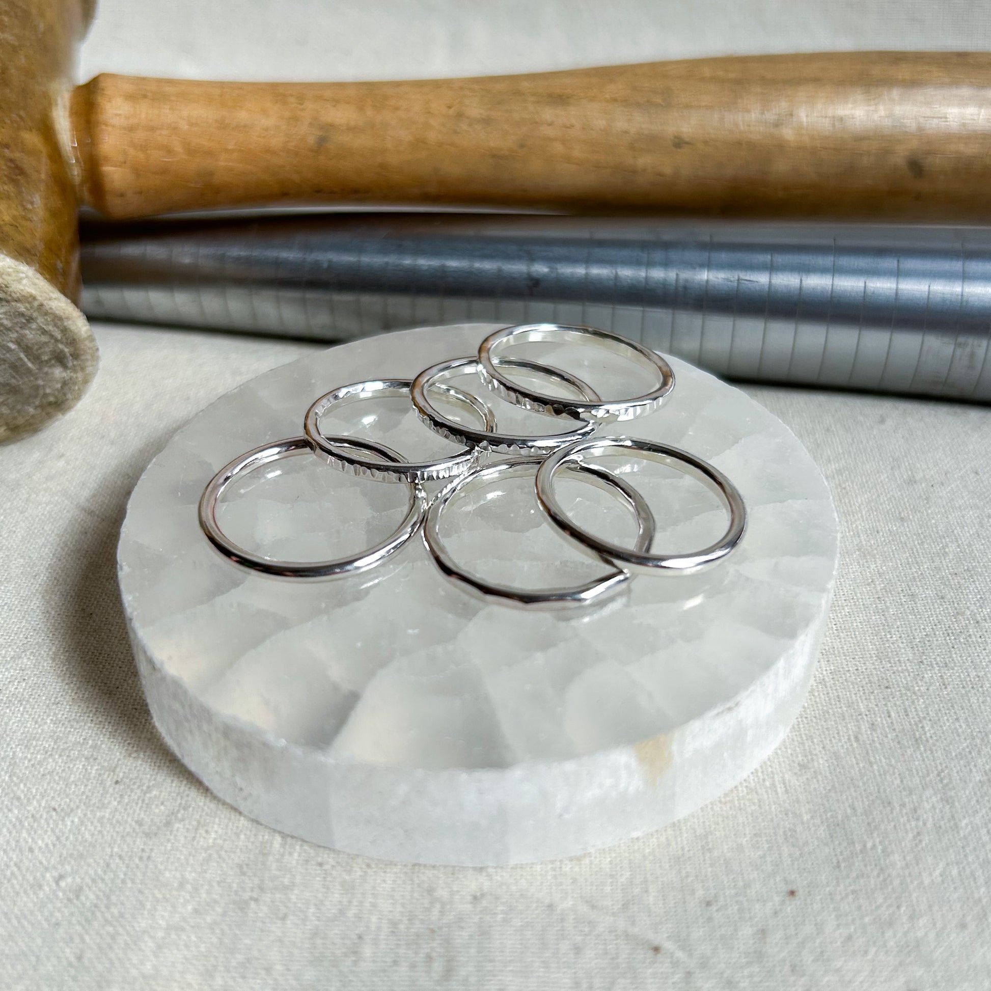 Five polished silver rings with textured, hammered edges are arranged on a round white marble coaster. In the background, a wooden mallet and a metal tube rest on a neutral fabric surface.