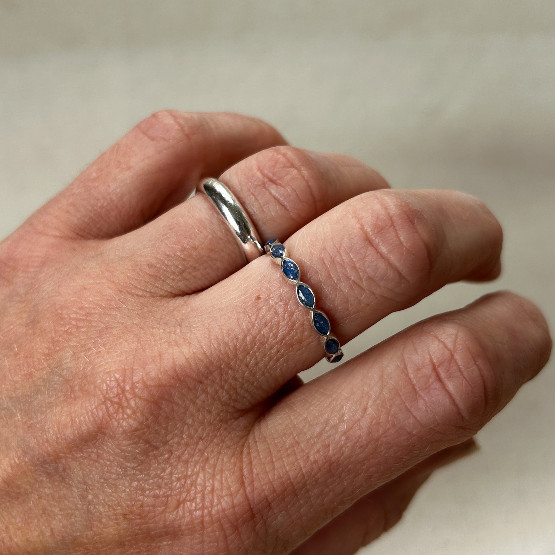 Hand wearing two rings, one silver and one with blue stones, on a neutral background