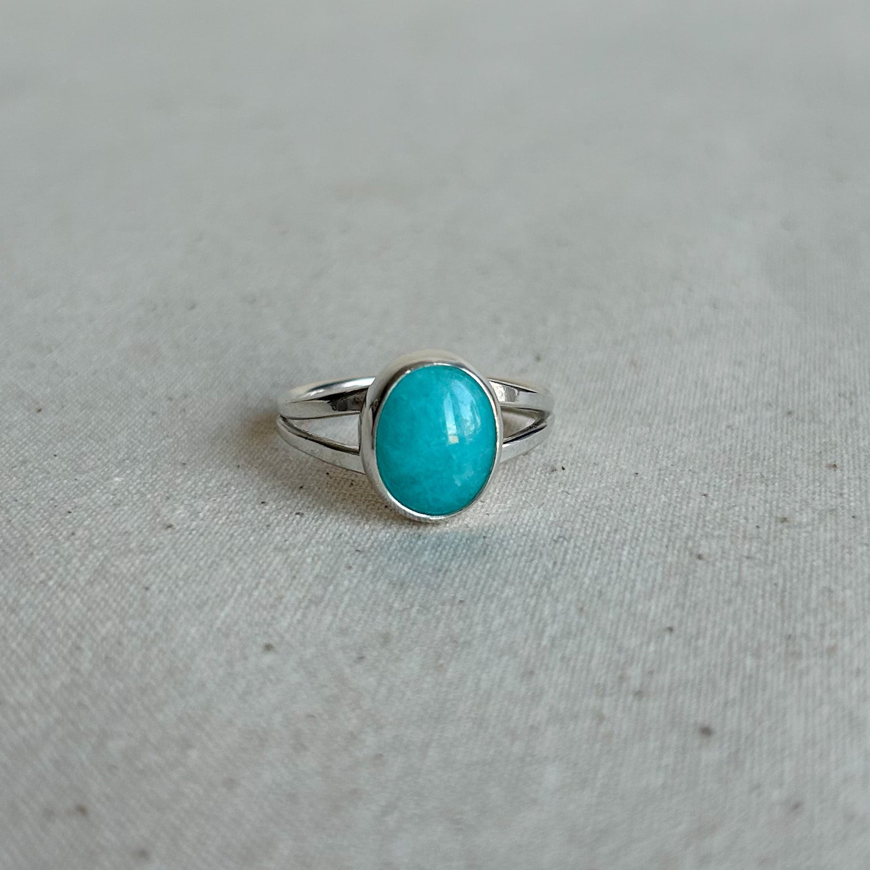 A silver ring with an oval turquoise cabochon stone, resting on a textured light gray fabric surface.