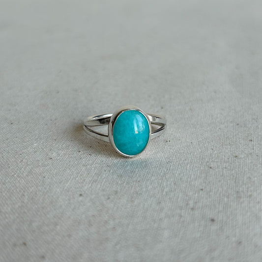 A silver ring with an oval turquoise cabochon stone, resting on a textured light gray fabric surface.