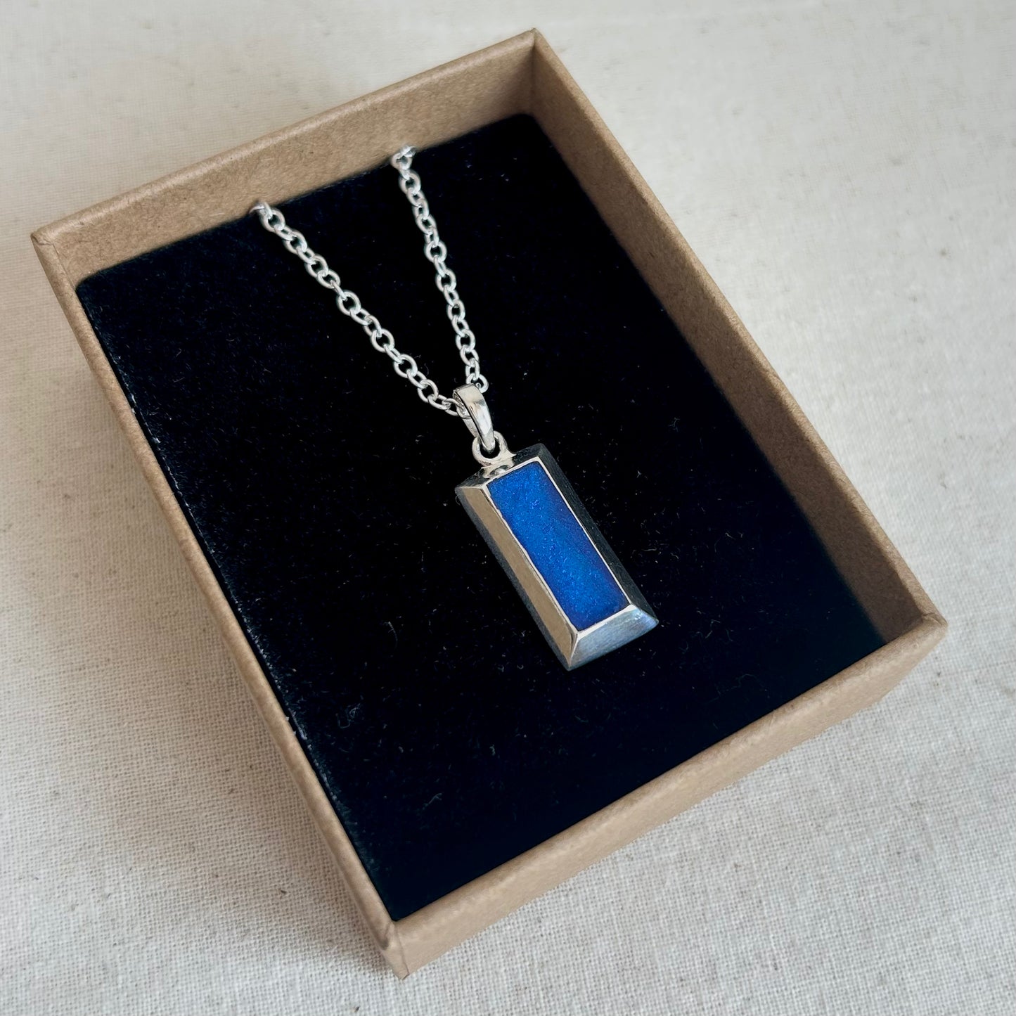 A silver-toned rectangular pendant necklace with a vibrant blue inlay, presented inside a brown cardboard gift box with a black velvet lining on a light beige fabric background.