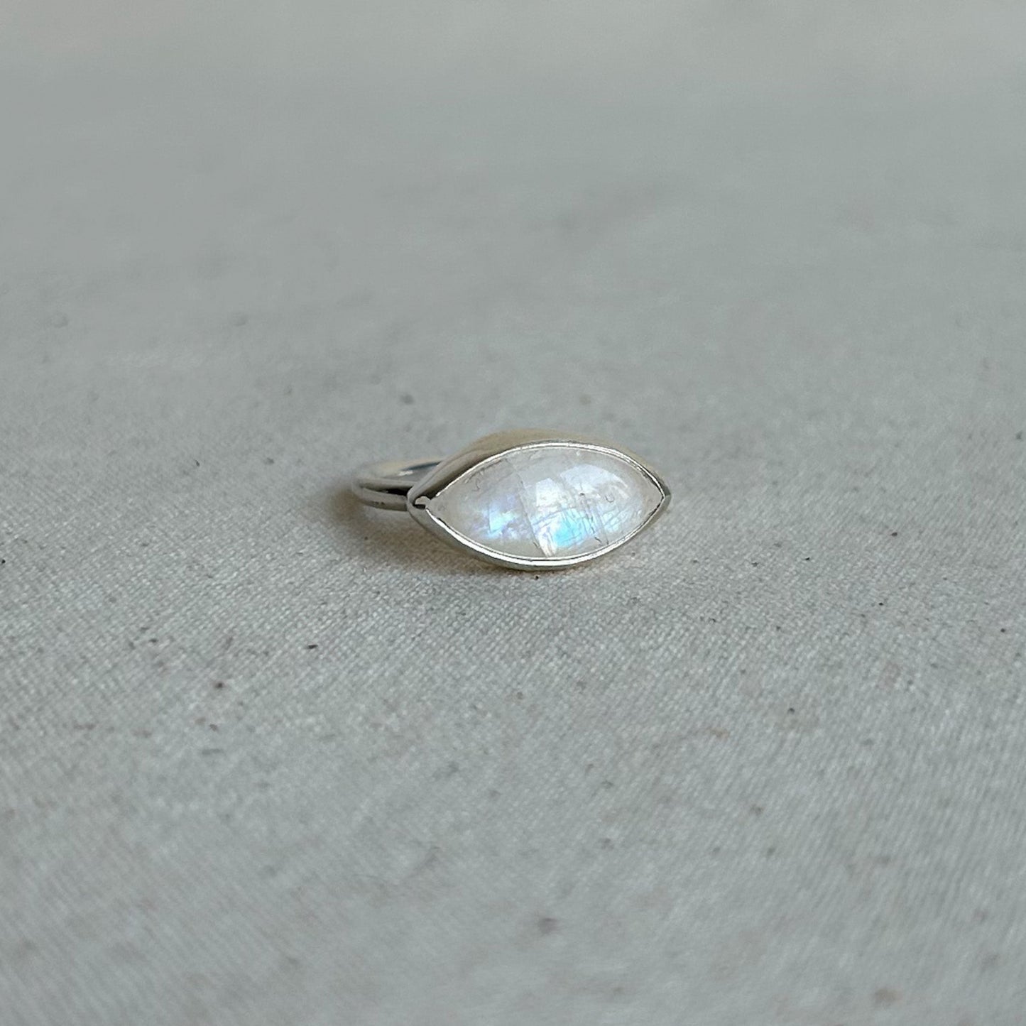 A silver ring featuring a marquise-cut iridescent moonstone gemstone, resting on a textured light gray fabric surface.