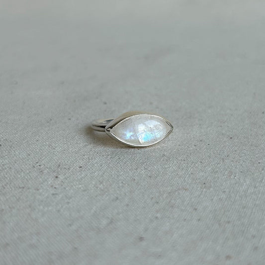 A silver ring featuring a marquise-cut iridescent moonstone gemstone, resting on a textured light gray fabric surface.