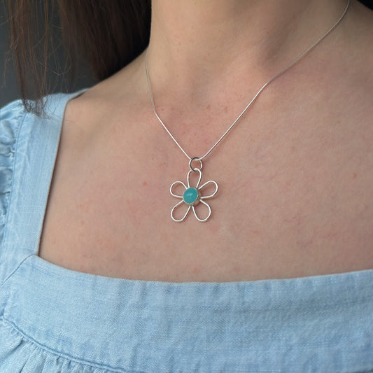 Necklace with a flower-shaped pendant featuring a turquoise stone, worn by a person in a light blue shirt.