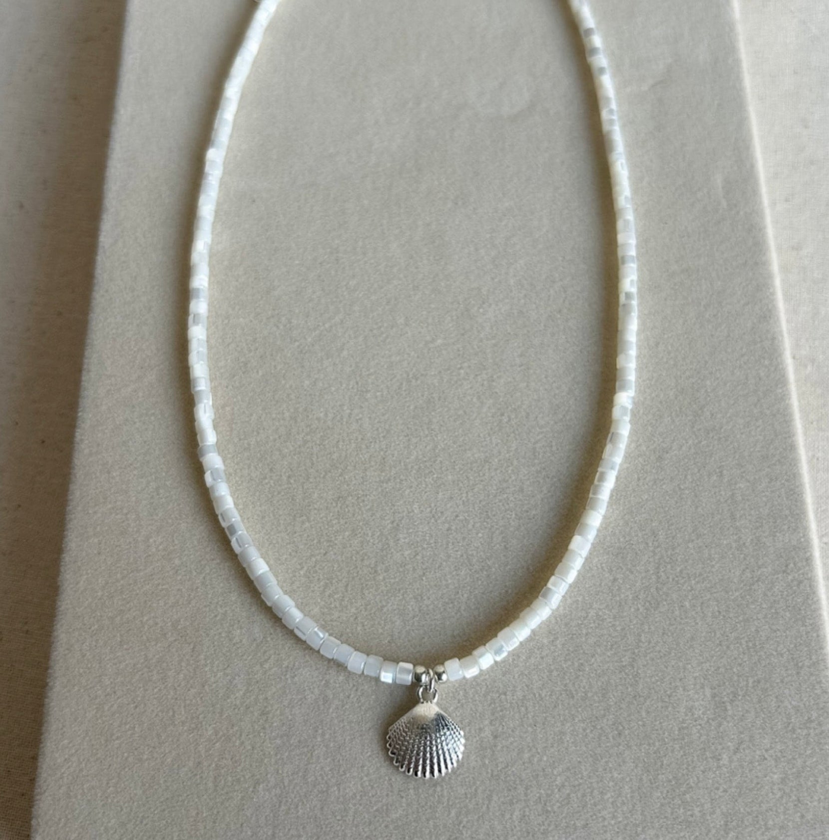 A delicate necklace made of small white beads with a silver seashell pendant, displayed on a neutral beige jewelry stand.