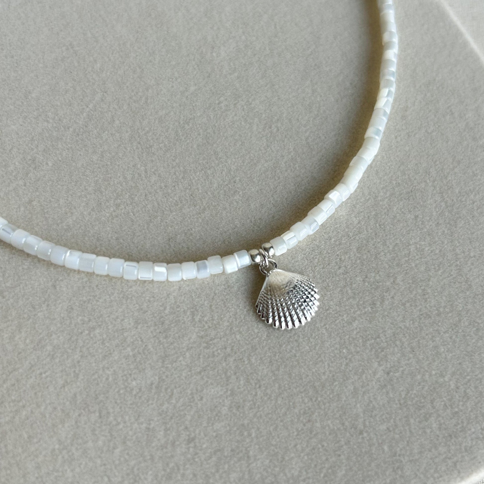 A delicate necklace made of small white rectangular beads with a small silver seashell pendant, displayed on a textured beige surface.