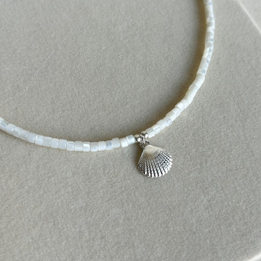A delicate necklace made of small white rectangular beads with a small silver seashell pendant, displayed on a textured beige surface.