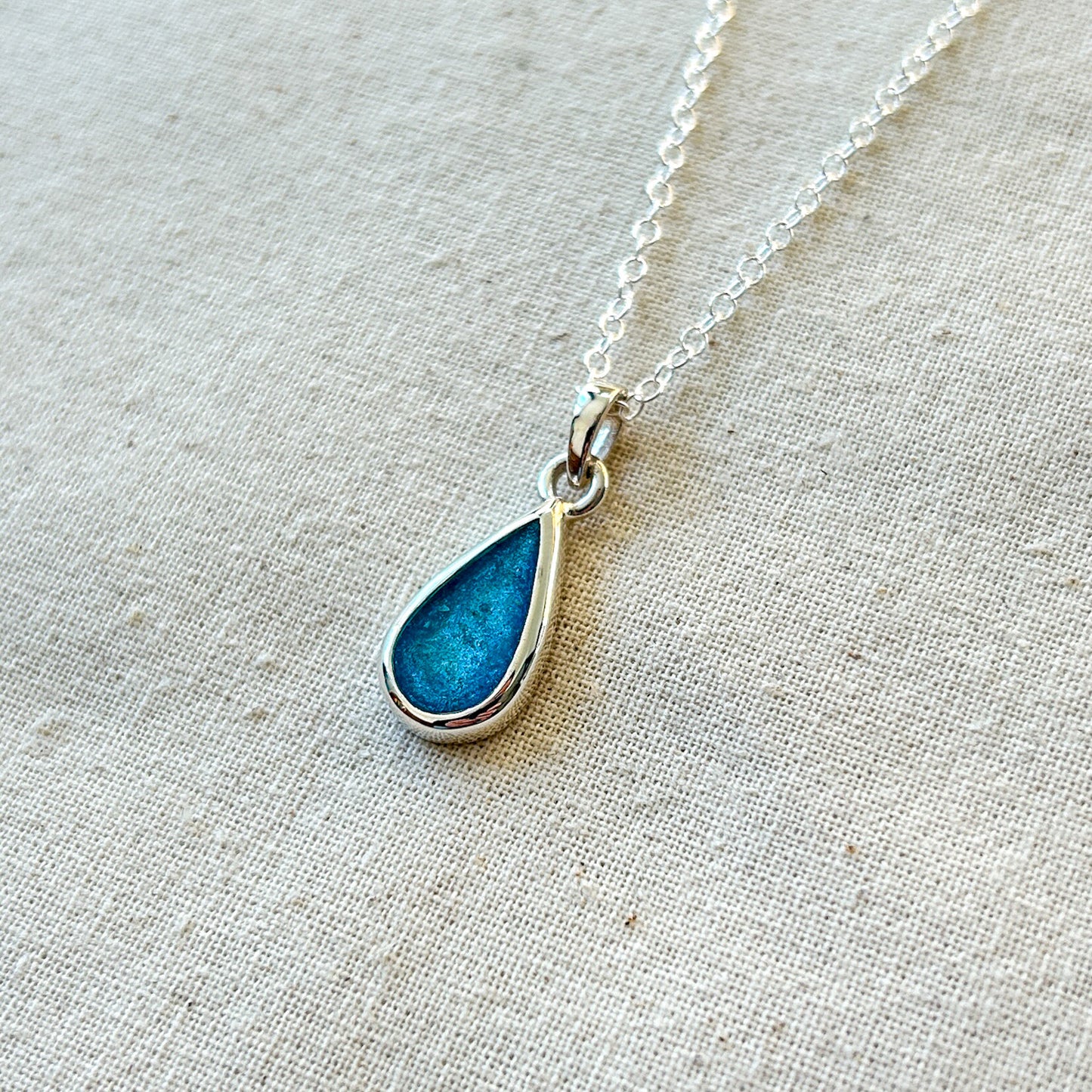 Teardrop-shaped blue pendant on a silver chain against a beige fabric background