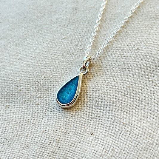 Teardrop-shaped blue pendant on a silver chain against a beige fabric background
