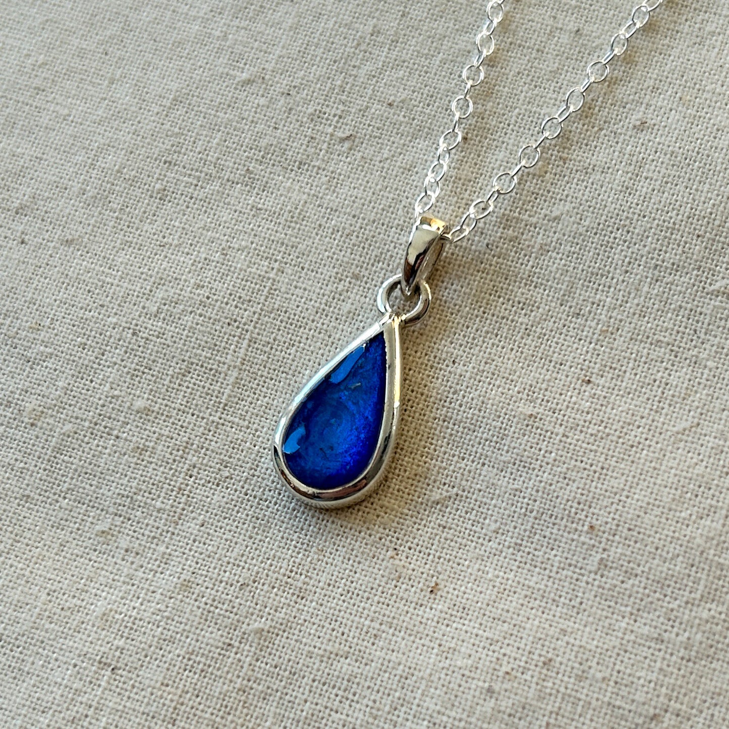 Teardrop-shaped blue pendant on a silver chain against a beige fabric background