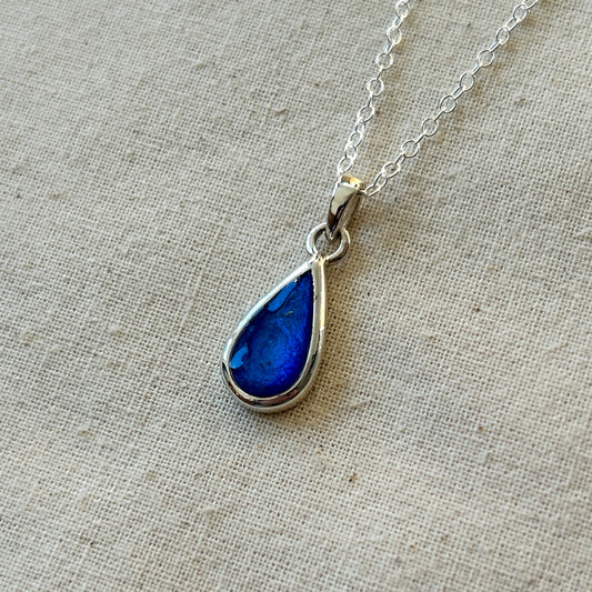 Teardrop-shaped blue pendant on a silver chain against a beige fabric background