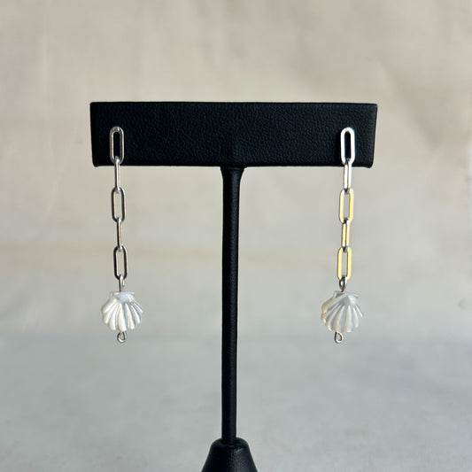 A pair of dangling earrings displayed on a black T-stand against a soft light gray background. Each earring has a silver paperclip chain leading to a white shell-shaped charm, with one earring also featuring a gold paperclip link.