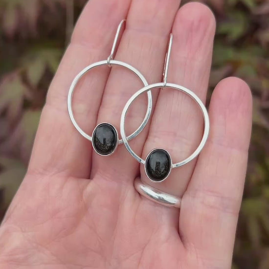 Silver and obsidian earrings held in hand against a natural blurred background